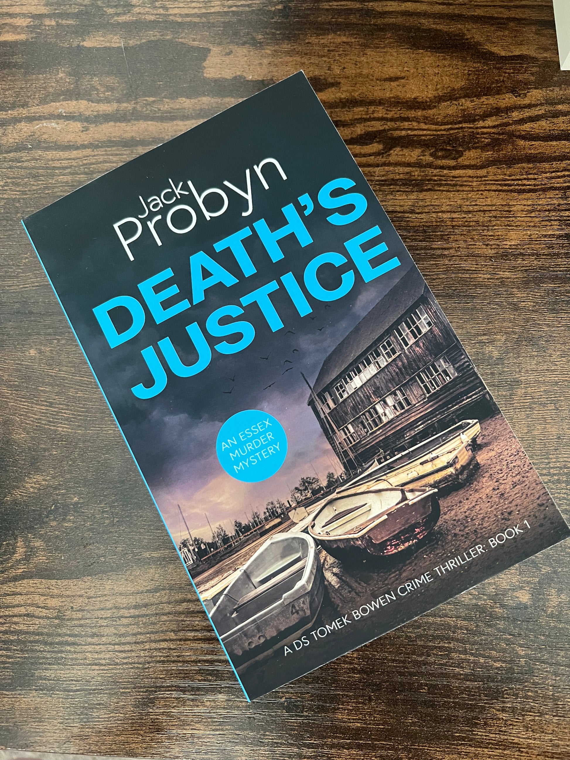 Book cover of Death's Justice, an Essex Murder Mystery by Jack Probyn. A gritty crime thriller featuring a weathered wooden boathouse on a muddy Essex shoreline at dusk. Book 1 in the DS Tomek Bowen series, perfect for fans of JD Kirk and JM Dalgliesh